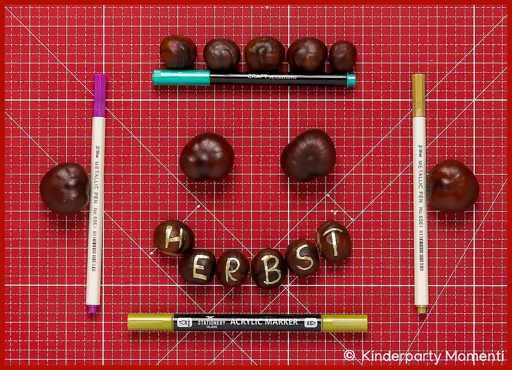 Kastanien liegen auf roter Schneidematte, einige mit den Buchstaben H E R B S T bemalt, umgeben von Acrylmarkern in verschiedenen Farben