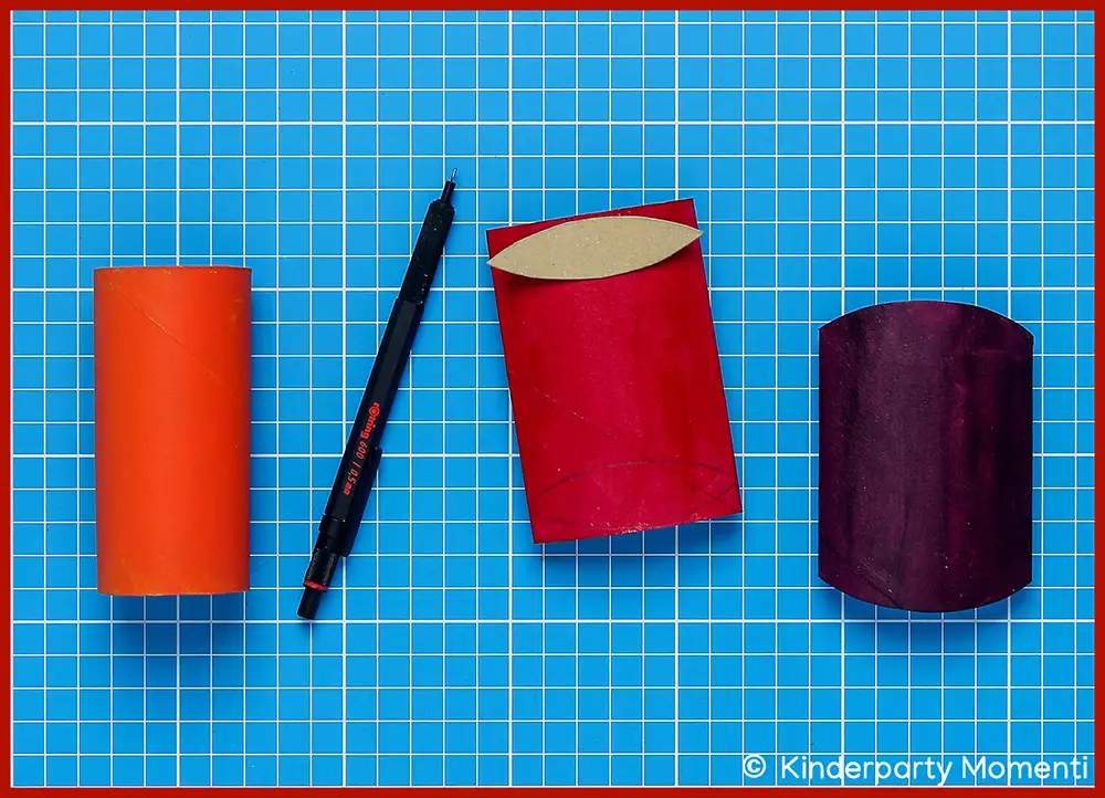 Kissenschachtel Vorlage auf Klopapierrollen übertragen Vier Gegenstände auf blauer Schneidematte: eine orangefarbene Klopapierrolle, ein schwarzer Stift, eine zusammengedrückte rote Klopapierrolle und eine zusammengedrückte lila Klopapierrolle