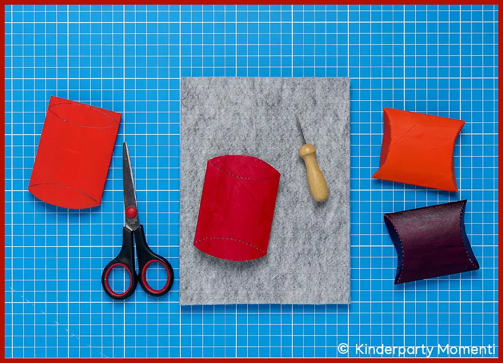 Falzlinie auf Toilettenpapaierrollen mit Prickelnadeln nachstechen Mehrere kleine, farbige Kissenschachteln aus Klopapierrollen, eine Schere, ein Ahle und eine graue Filzunterlage auf blauer Schneidematte