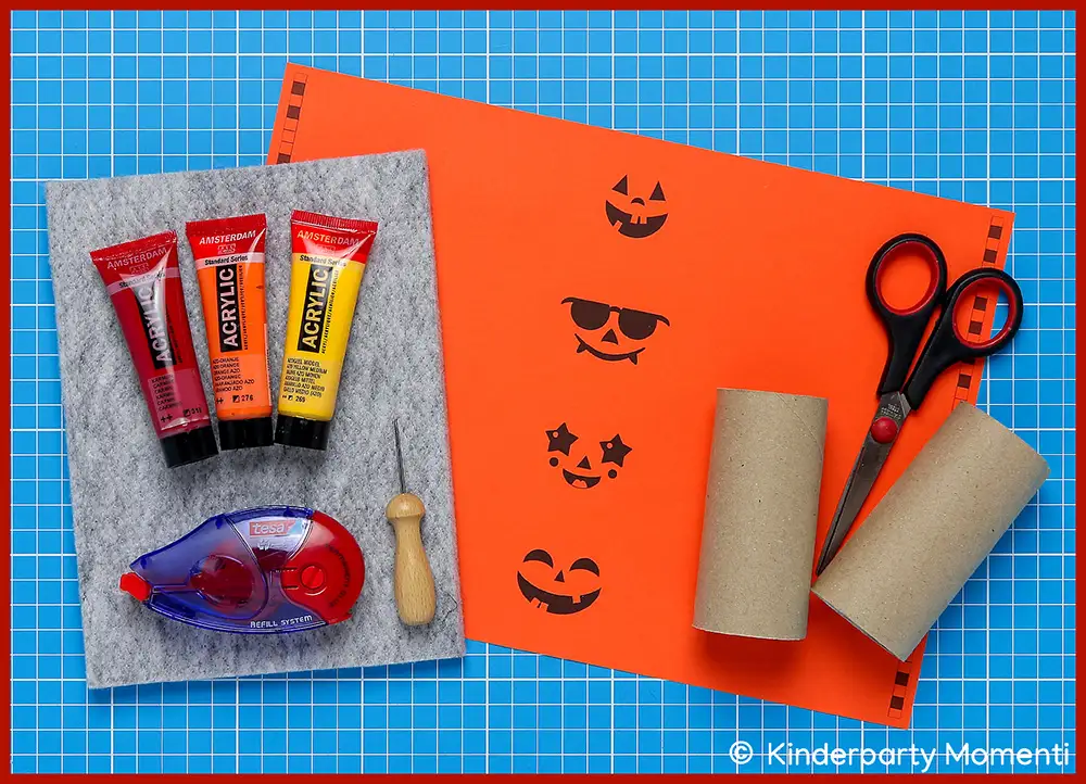 Material zum Halloween Kissenschachteln aus Klopapierrollen basteln Bastelmaterialien auf blauer Schneidematte mit Filzplatte, Acrylfarben, Kleberoller, Prickelnadel, orangenem Papier mit Kürbisgesichtern, Schere und zwei leeren Klopapierrollen