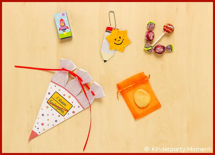 Kleine Schultüte mit Punkten, rotem Band und Aufkleber 'Schule Zukunft', daneben Schlüsselanhänger mit Perlen, Bonbons, Lutscher, kleiner orangener Beutel und Kartenspiel.