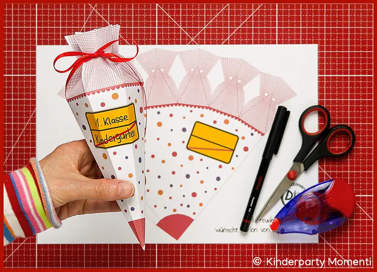 Hand hält eine gebastelte Zuckertüte mit Aufklebern '1. Klasse' und 'Kindergarten' vor Bastelvorlagen, Schere, Stift und Kleber auf roter Bastelmatte.