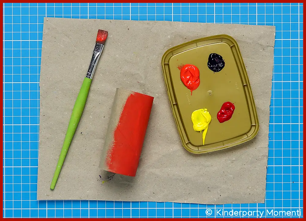 Klopapierrollen mit Halloween Farben bemalen Pinsel mit roter Farbe, halb bemalte Klopapierrolle und Farbpalette mit roten, gelben und lila Farben auf brauner Papierunterlage auf blauer Schneidematte