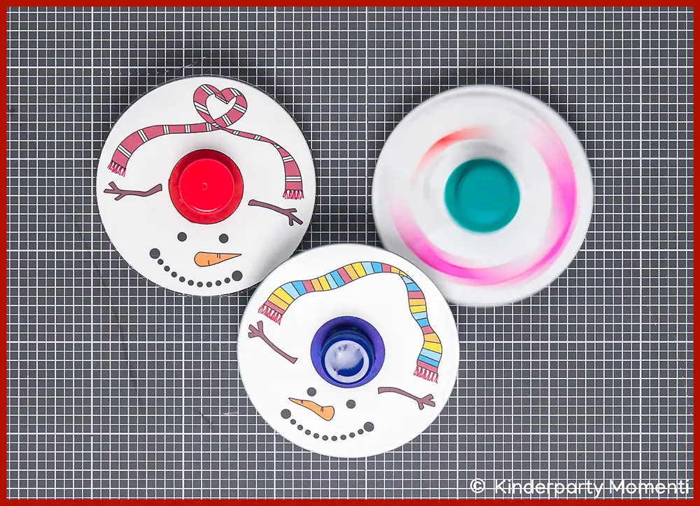 DIY Kreisel Spiele selber basteln mit CDs Drei selbstgebastelte Kreisel aus CDs mit aufgemalten Schneemann-Gesichtern und bunten Schals auf grauer Bastelmatte, einer der Kreisel ist in Bewegung