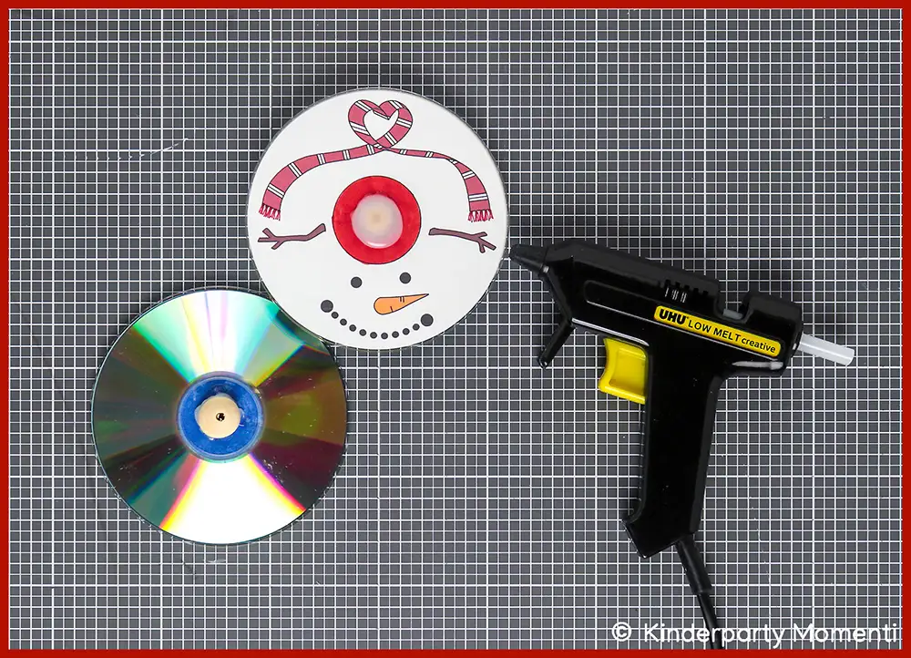 02 Kreisel Basteln aus CDs - Holzkugel als Drehpunkt unter die CD kleben Zwei CDs, eine mit Schneemann-Gesicht und Mütze bemalt, daneben eine Heißklebepistole auf grauer Bastelmatte