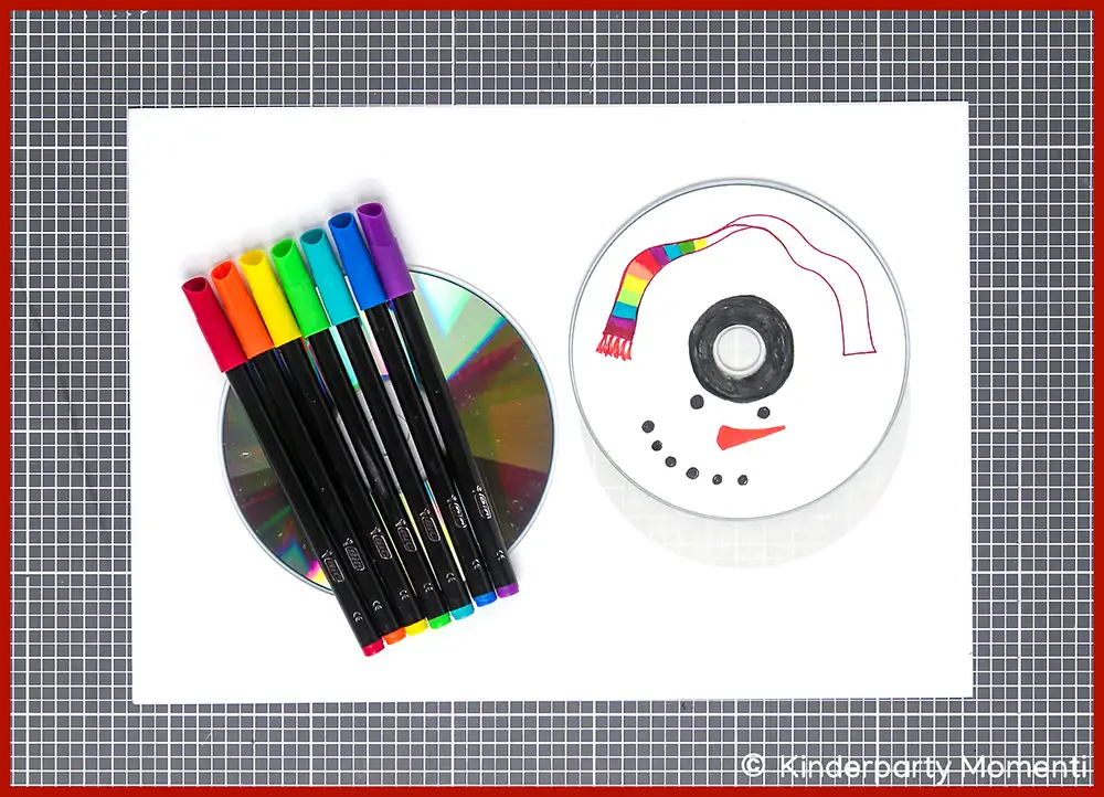 01 Kreisel Basteln aus CDs - Etiketten für CDs mit Stiften bemalen Zwei CDs auf grauer Bastelmatte, eine mit bunten Filzstiften darauf, die andere mit einem Schneemanngesicht und einem bunten Schal bemalt