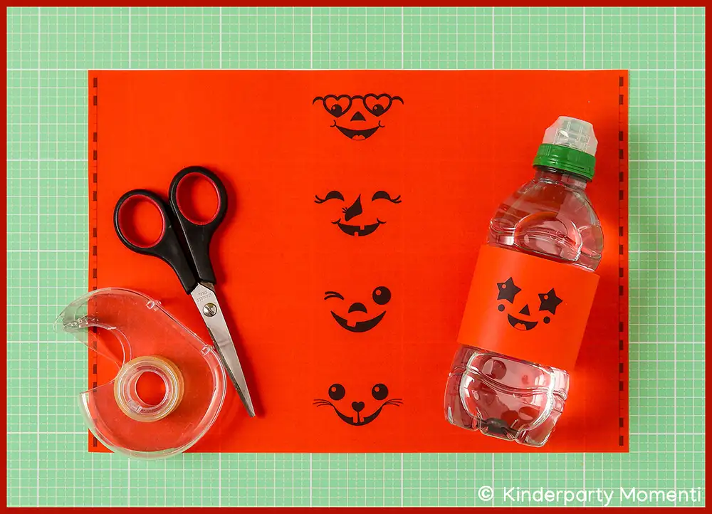 Ausgedruckte Flaschenetiketten für Halloweenpartys Schere, Klebeband und eine Flasche mit orangefarbenem Etikett mit drei lachenden Halloween-Kürbisgesichtern auf rotem Bastelpapier