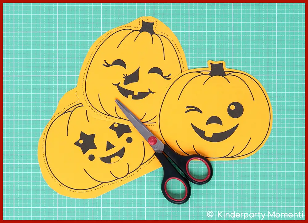 Halloween Girlanden basteln mit lusitgen Kürbisgesichtern Auf einer Bastelmatte liegen drei gelbe Kürbisse aus Papier mit lustigen Kürbisgesichtern und eine Schere.