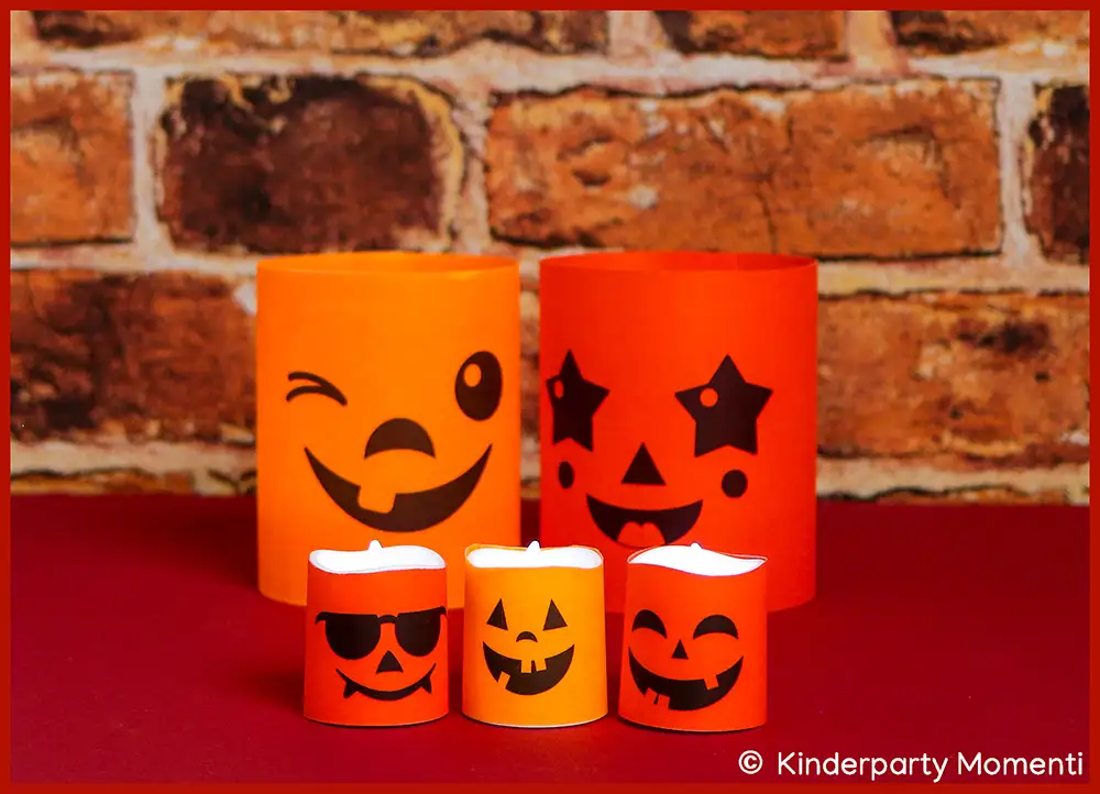 Orangene und rote Windlichter und LED Teelichter mit lachenden Halloween Kürbisgesichtern auf roter Tischdecke und vor Backsteinwand