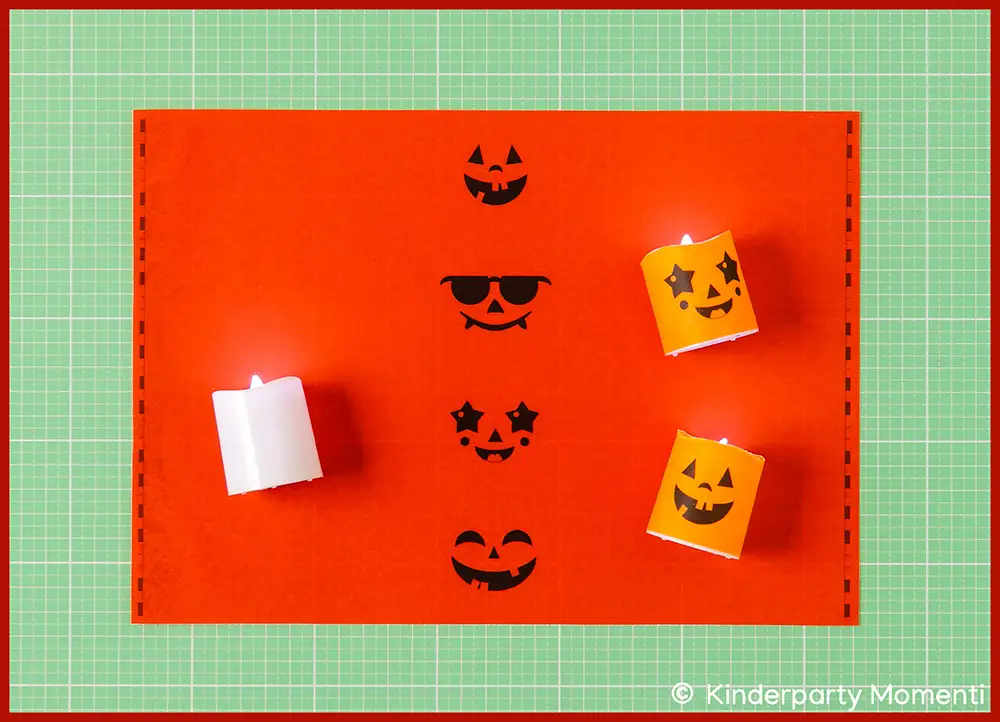 Zwei orangefarbene Kerzen mit Halloween Gesichtern und eine weiße Kerze auf roter Papier Vorlage mit schwarzen Halloween Kürbisgesichtern