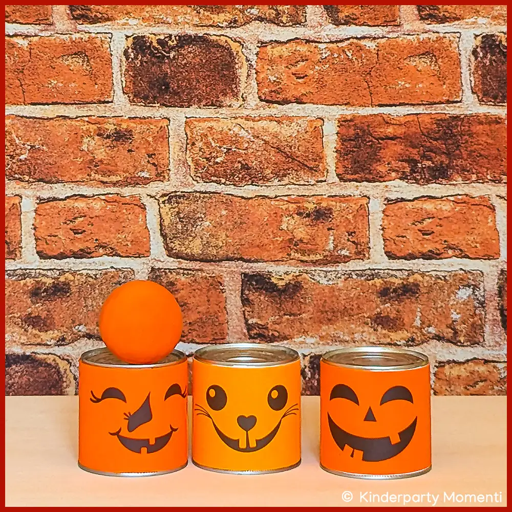 DIY Dosenwerfen für Halloween nach dem Wurf Drei beim Dosenwerfen stehengebliebene orangefarbene Dosen mit Halloween-Gesichtern, eine Dose mit aufgesetztem orangefarbenem Ball, vor roter Backsteinwand