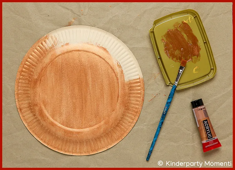 Pappteller, der mit brauner Farbe bemalt wird, daneben Pinsel und Acrylfarben-Tube auf braunem Untergrund