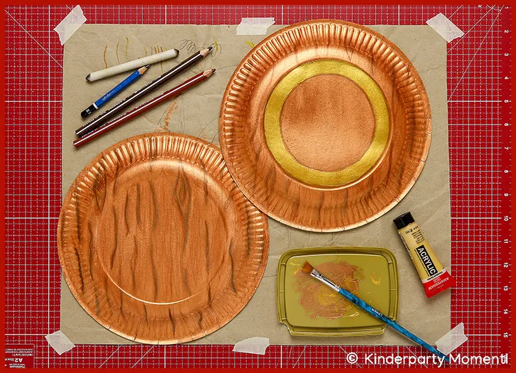 Zwei bemalte Pappteller mit braunen und goldenen Farbtönen, daneben Pinsel, Acrylfarbe, Buntstifte und ein Stift auf roter Bastelmatte