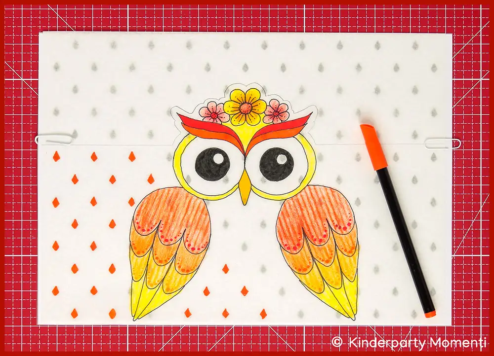 Bunte Zeichnung einer Eule mit großen Augen, roten und gelben Flügeln, verziert mit Blumen auf dem Kopf, daneben ein oranger  Filzstift zum Abmalen des Mustern, die Eulen Vorlage liegt auf einer roten Bastelmatte