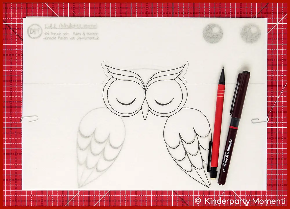 Laminiertes Butterbrotpapier liegt auf einer Vorlage zum Basteln einer Eulen Laterne, darauf zwei Stifte zum nachmalen der Vorlage, alles liegt auf einer roten Bastelmatte