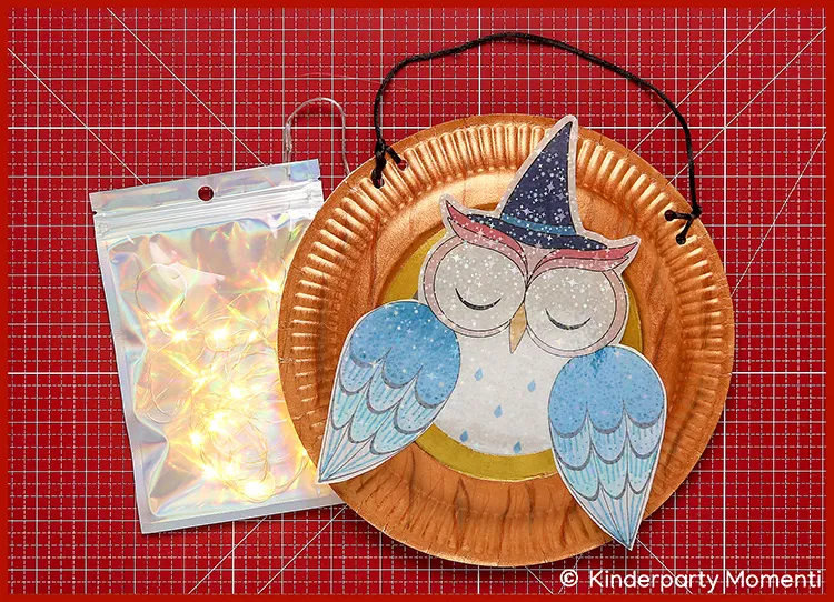 Pappteller mit einer Eule aus Papier, die eine Hexenhut trägt, daneben eine kleine Lichterkette in einer durchsichtigen Verpackung