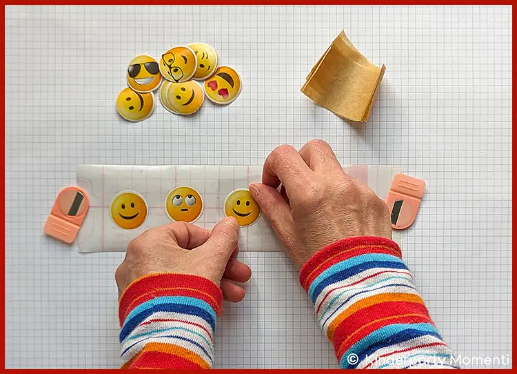 Hände mit bunt gestreiften Ärmeln kleben gelbe Emoji-Sticker auf eine weiße Klebefläche auf kariertem Untergrund