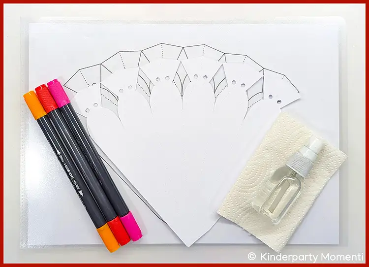 Schultüten-Vorlage aus Papier mit Markierungen, daneben bunte Filzstifte und eine kleine Sprühflasche auf einem Papiertuch