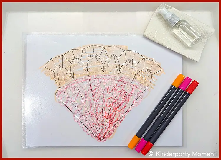 Teilweise ausgemalte Schultütenvorlage auf Papier neben vier Filzstiften in Orange, Pink, Rot und Schwarz sowie einer kleinen Sprühflasche auf einem weißen Tuch