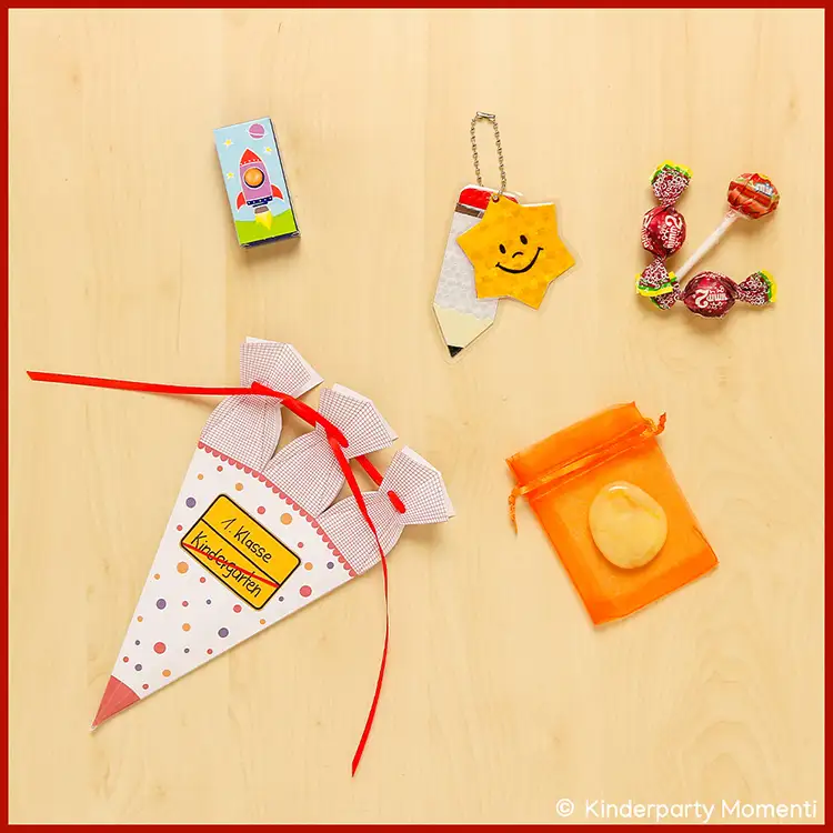 Kleine gebastelte Schultüte mit rotem Band und Aufschrift '1. Klasse Kindergarten' neben einem Reflektor in Form eines Bleistifts mit gelbem Stern und einem orangefarbenen Organzasäckchen mit einem Stein sowie Süßigkeiten.
