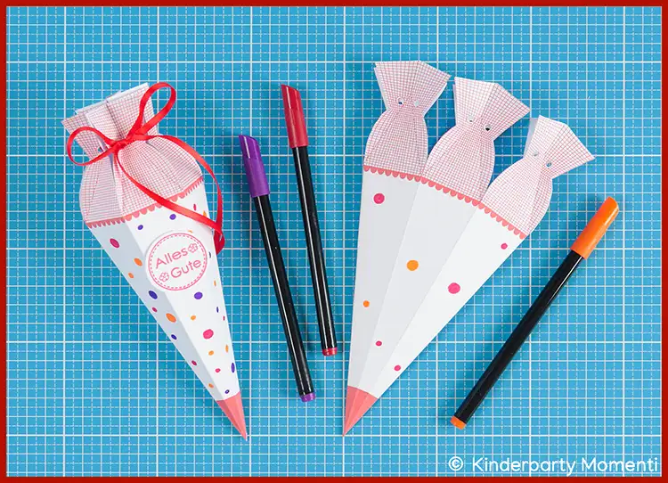 Mini Schultüten selbst basteln und bemalen Zwei weiße Papier Schultüten mit roten Spitzen und bunten Punkten, eine mit roter Schleife und der Aufschrift 'Alles Gute', daneben drei Filzstifte in Rot, Lila und Orange auf blauer Bastelmatte