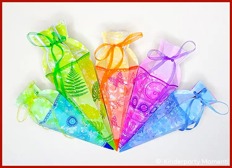 Fünf bunte, transparente Mini-Schultüten mit verschiedenen Stempelmotiven und Schleifenbändern, angeordnet fächerförmig auf weißem Hintergrund