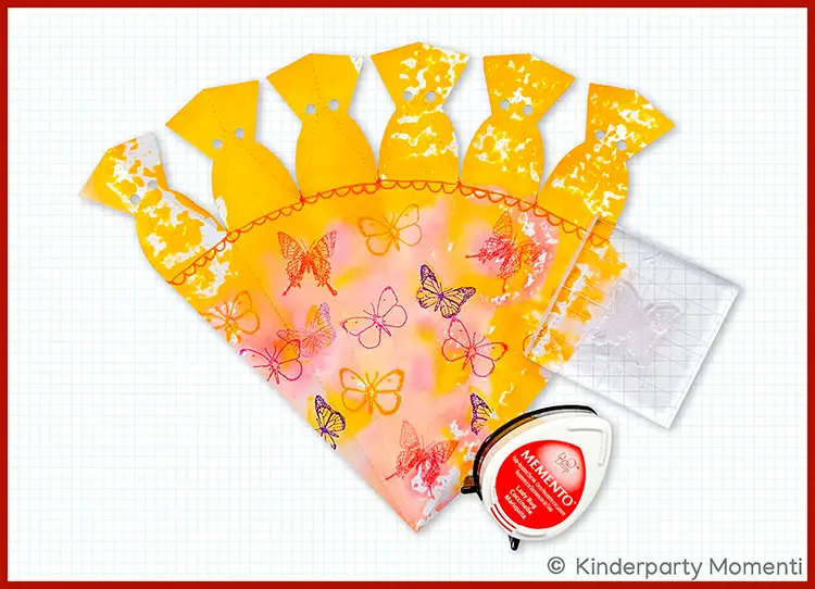 Vorlage für gelb-orange Schultüte aus Papier mit aufgedruckten Schmetterlingen, daneben ein rotes Stempelkissen und ein transparentes Stempelgitter
