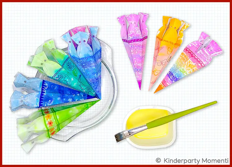 Mehrere bunte, handbemalte Mini-Schultüten aus Papier, die auf einem weißen Untergrund liegen, daneben ein Pinsel und eine kleine Schale mit gelber Farbe