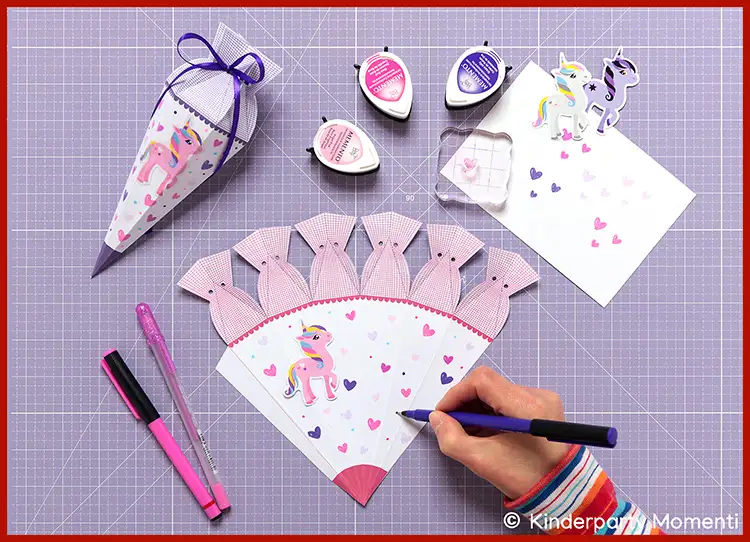 Hand malt mit lila Stift ein Einhorn und kleine Herzen auf eine halbkreisförmige Papiervorlage, um eine Zuckertüte zu gestalten, umgeben von Bastelmaterialien und fertiger Zuckertüte mit Einhornmotiv