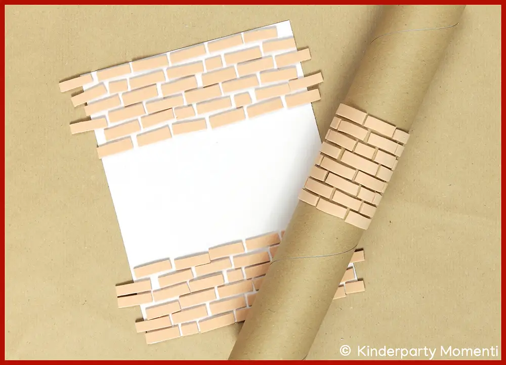 DIY Moosgummi Stempelrolle aus brauner Papprolle mit aufgeklebten, schmalen, rechteckigen Moosgummistreifen, die ein Mauerwerk-Muster bilden, liegt auf braunem Papier neben einem weißen Blatt mit ähnlichem Muster.