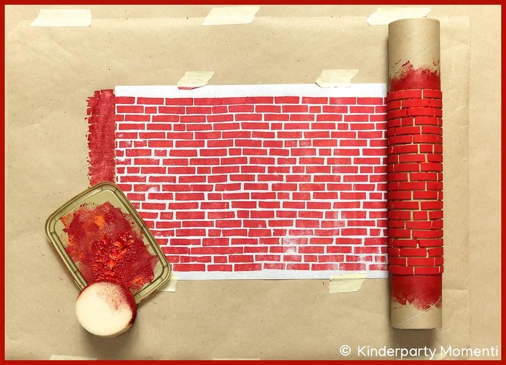 Stempel Rolle aus Moosgummi mit Ziegelmuster, daneben ein mit roter Farbe bedeckter Schwamm und ein weißes Blatt mit rotem Ziegelmuster auf braunem Papier.
