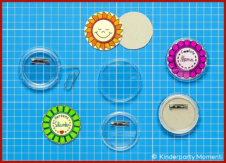 Runde selbstgemachte Ansteckbuttons mit Sonnen- und Blumenmotiven auf blauem Karopapier, teils mit Sicherheitsnadel-Rückseite