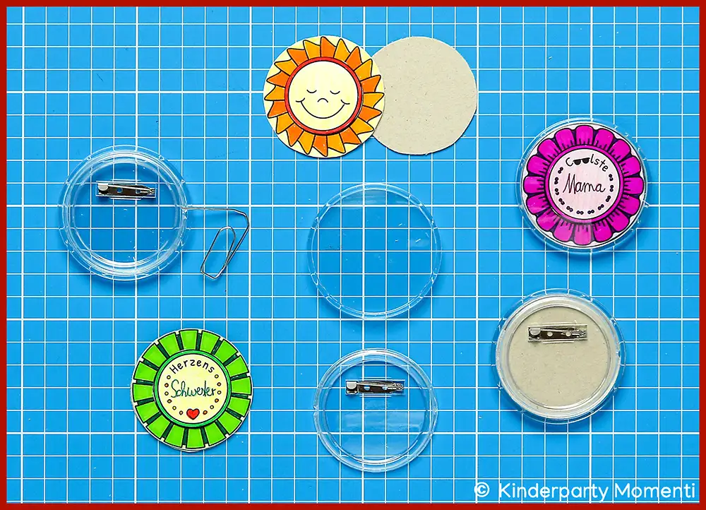 Runde selbstgemachte Ansteckbuttons mit Sonnen- und Blumenmotiven auf blauem Karopapier, teils mit Sicherheitsnadel-Rückseite
