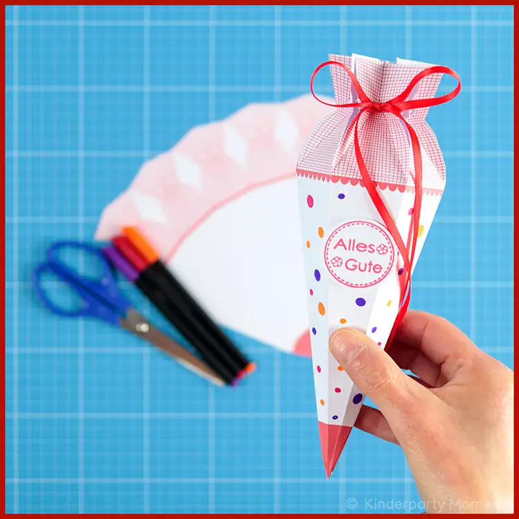 Eine Hand hält eine kleine selbstgebastelte Schultüte auf der 'Alles Gute' steht. Im Hintergrund ist die Schultüten Vorlage und Bastelwerkzeug zu sehen.