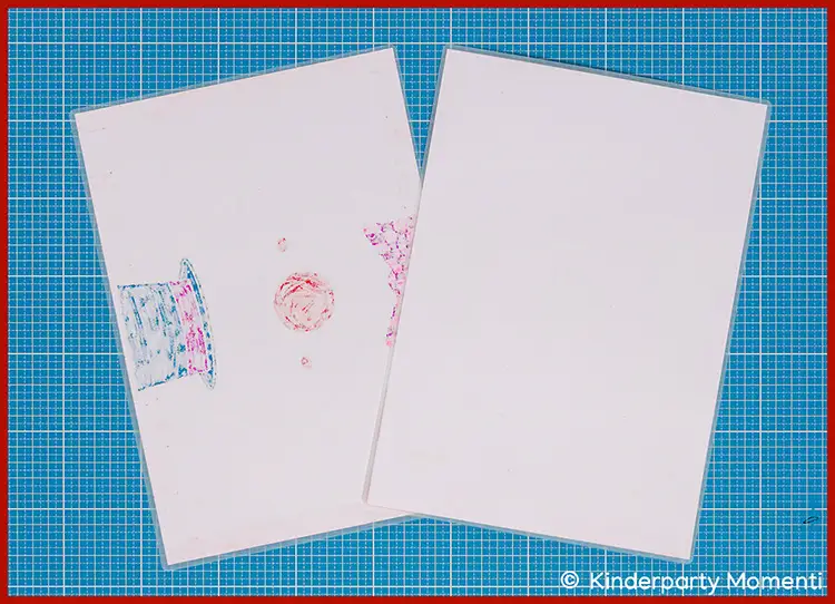 Zwei weiße Laminierte Papierbögen auf blauem Schneideunterlage mit Raster, einer mit teilweise sichtbarer bunter Zeichnung