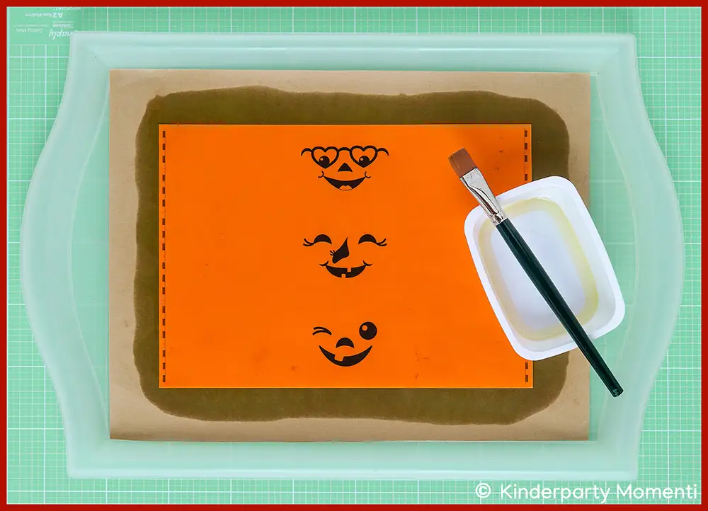 Ausgedruckte Vorlage für Halloween Flaschenetiketten mit Öl transparent machen Orangefarbene Papier Vorlage mit drei lachenden Kürbisgesichtern, daneben ein Pinsel und eine weiße Schale mit Öl um das Papier durchscheinend zu machen