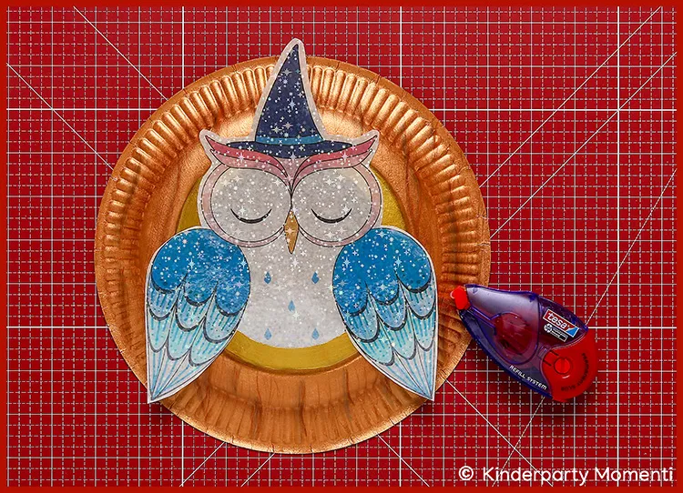 Pappteller mit aufgeklebter Eulenlaterne aus Papier und Kleberoller auf roter Bastelmatte