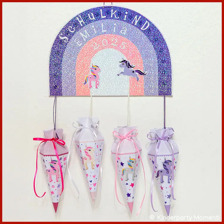 Vier Schultüten mit Einhornmotiven und Schleifen in Rosa, Weiß und Lila hängen unter einem Regenbogen mit der Aufschrift 'Schulkind Emil 2025'