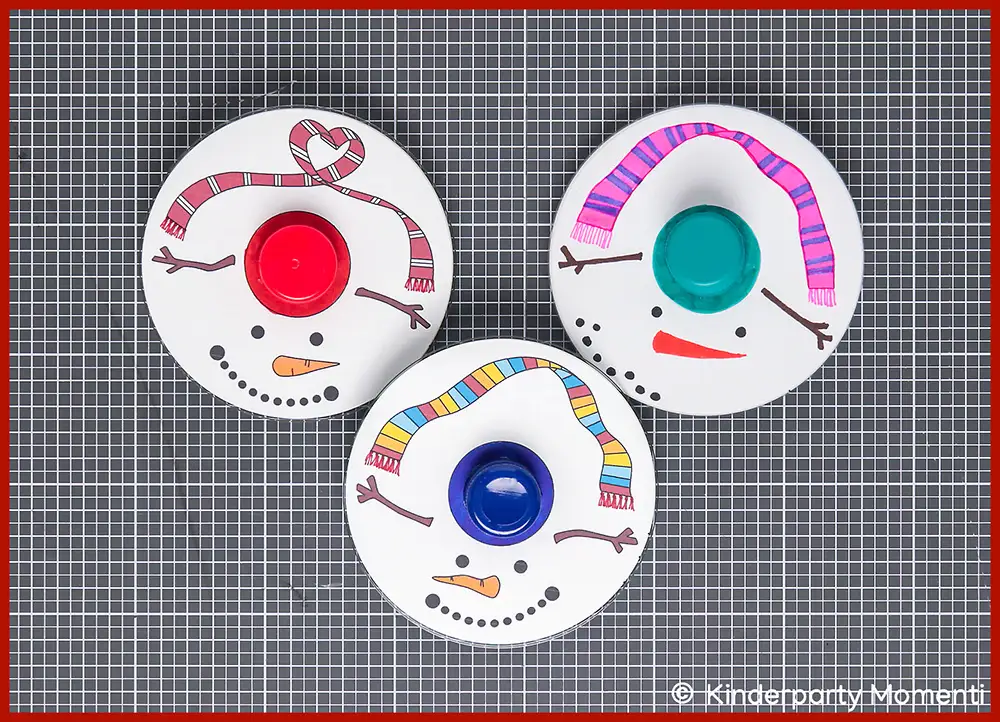 Schneemann Kreisel selber basteln aus CDs Drei selbstgebastelte Schneemann-Kreisel aus CDs mit bunten Mützen, Schals und Gesichtern aus Punkten und Strichen auf grauer Bastelmatte