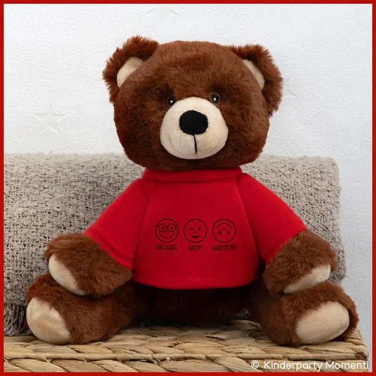 Brauner Teddybär mit rotem Pullover und aufgedruckten drei Gesichtern sitzt auf geflochtenem Untergrund vor hellgrauer Wand