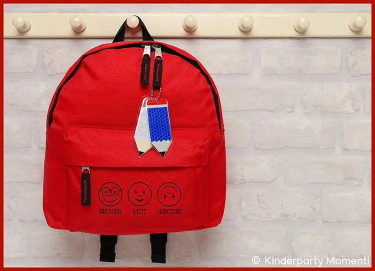 Kleiner Rucksack für Grundschulkinder Roter Kinderrucksack mit drei aufgedruckten Gesichtern und den Worten 'MUT', 'MUT', 'ABENTEUER' vor heller Ziegelsteinwand
