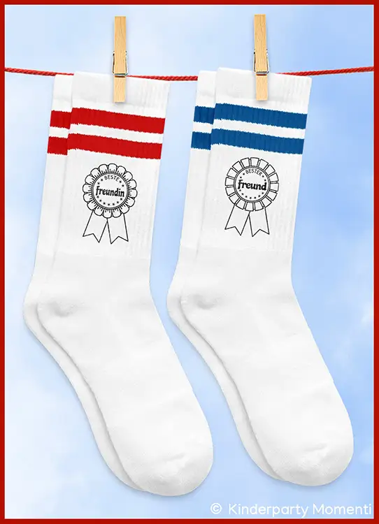 Zwei weiße Socken mit je zwei Streifen oben, eine mit roten Streifen und Aufdruck 'Beste Freundin', die andere mit blauen Streifen und Aufdruck 'Bester Freund'