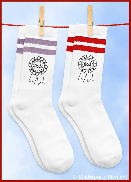 Zwei weiße Socken mit je zwei farbigen Streifen oben und einem aufgedruckten Abzeichen mit den Worten 'Beste Godi' und 'Bester Göd' hängen an einer Wäscheleine mit Wäscheklammern