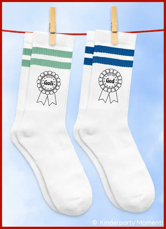 Zwei weiße Socken mit je zwei farbigen Streifen oben und einem aufgedruckten Rosettenmotiv mit den Worten 'Bester Goli' und 'Bester Ged' hängen an einer Wäscheleine mit Holzklammern
