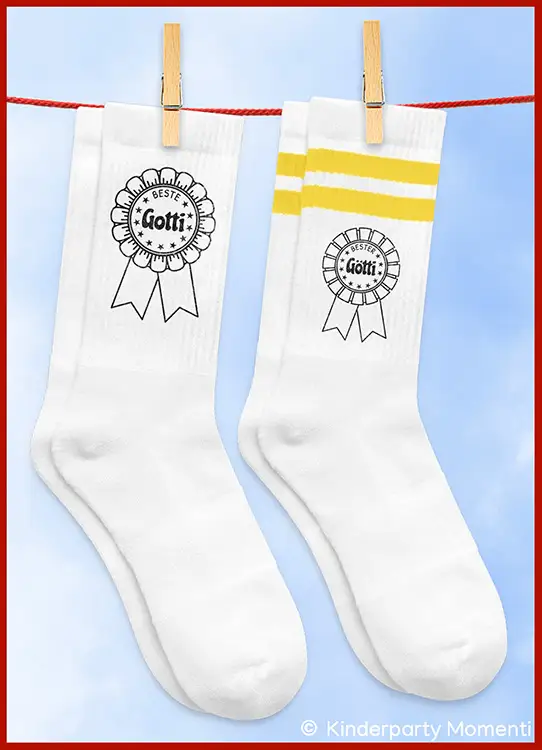 Zwei weiße Socken mit Aufdruck, eine mit gelben Streifenmuster und der Schrift 'Beste Gotti', die andere mit der Schrift 'Bester Götti', beide an einer Wäscheleine befestigt