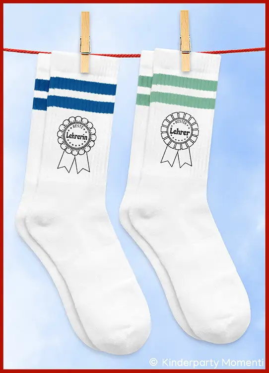 Zwei weiße Socken mit farbigen Streifen oben, eine mit blauen Streifen und Aufdruck 'Beste Lehrerin', die andere mit grünen Streifen und Aufdruck 'Bester Lehrer', an einer Wäscheleine mit Wäscheklammern hängend