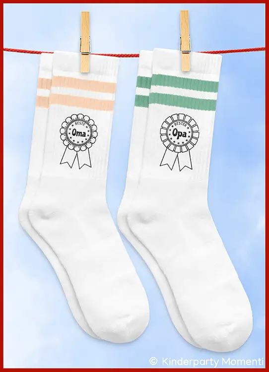 Zwei weiße Socken mit je zwei Streifen oben, eine mit hellroten Streifen und der Aufschrift 'Beste Oma', die andere mit grünen Streifen und der Aufschrift 'Bester Opa', an einer Wäscheleine mit Wäscheklammern aufgehängt