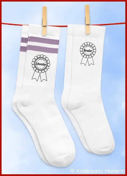 Zwei weiße Socken an einer Wäscheleine, eine mit zwei lilafarbenen Streifen und der Aufschrift 'Beste Schwester', die andere mit der Aufschrift 'Bester Bruder'