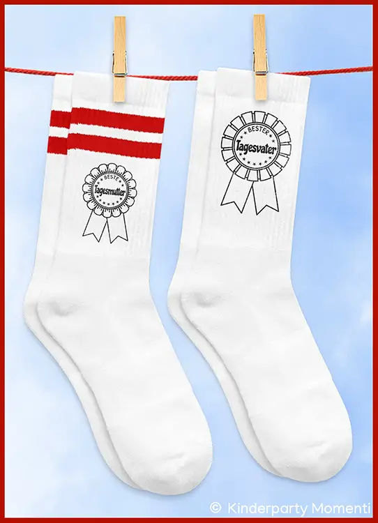 Zwei weiße Socken an einer Wäscheleine, eine mit roten Streifen und der Aufschrift 'Beste Tagesmutter', die andere mit der Aufschrift 'Bester Tagesvater'
