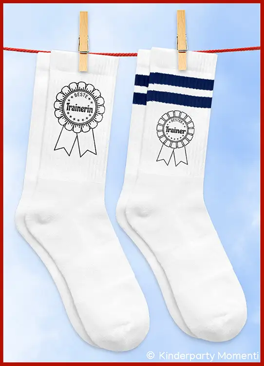 Zwei weiße Socken mit Aufdruck einer Siegerrosette, eine mit dem Text 'Beste Trainerin', die andere mit 'Bester Trainer' und zwei dunklen Streifen am oberen Rand